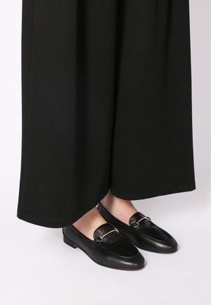 Schwarze Lederloafers mit abgerundetem Zehenbereich, goldfarbenen Hardware-Akzenten und einem niedrigen Absatz, kombiniert mit einem fließenden schwarzen Kleidungsstück.