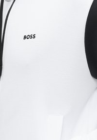 Λευκό υφασμάτινο ύφασμα με μαύρο περίγραμμα και τη λέξη «BOSS» κεντημένη μαύρη στην πάνω περιοχή του στήθους.