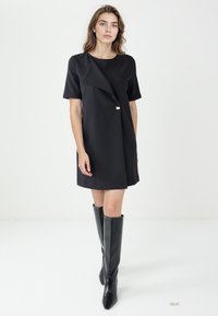 Vestito nero con maniche corte, davanti asimmetrico e bottone decorativo dorato. Indossato con stivali neri fino al ginocchio, tessuto liscio e taglio sartoriale.