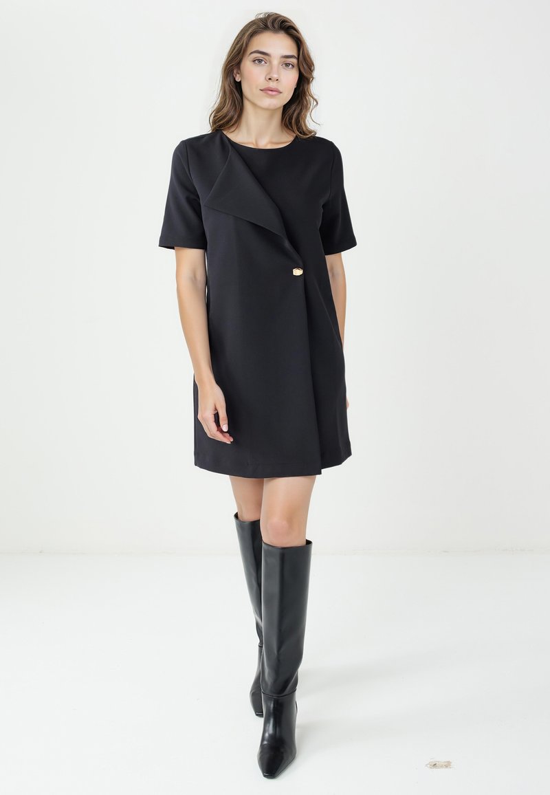 Vestito nero con maniche corte, davanti asimmetrico e bottone decorativo dorato. Indossato con stivali neri fino al ginocchio, tessuto liscio e taglio sartoriale.