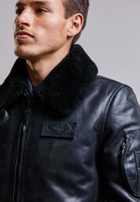 Blouson en cuir noir avec un col en shearling noir doux, fermeture éclair sur le devant et un patch de marque en relief sur la poitrine. Caractérisé par une poche zippée sur le bras.