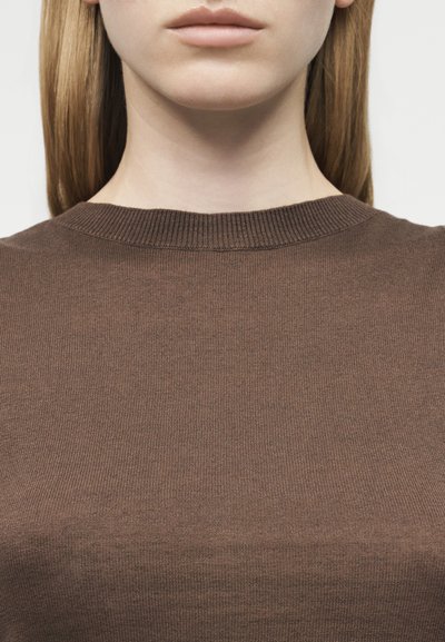 Κοντινό πλάνο του κάτω μέρους του προσώπου ενός ατόμου και του λαιμού που φοράει καφέ πουλόβερ με στρογγυλή λαιμόκοψη, με φόντο ένα απλό ανοιχτόχρωμο φόντο.