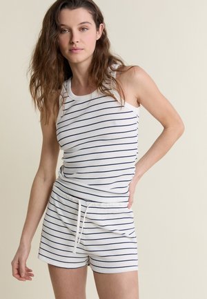 Joven con cabello castaño ondulado que lleva una camiseta sin mangas blanca de canalé y pantalones cortos a juego con cordón, con finas rayas horizontales azul marino.