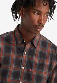 Camisa de cuadros para hombre en tonos oscuros con rayas naranjas y azules. Cuenta con un cuello abotonado y un pequeño logo en el bolsillo del pecho.