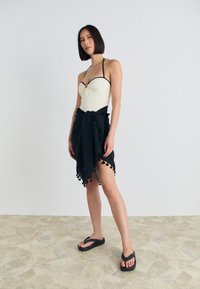 Krämfärgad baddräkt med svarta detaljer, lager av svart sarong med tofsar, och svarta flip-flops. Stående på en strukturerad beige golv.