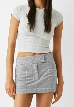 Femme portant un crop top gris clair ajusté et une mini-jupe à carreaux grise taille haute avec une ceinture asymétrique.