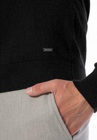 Czarny sweter z dzianiny z prążkowanymi mankietami i dołem, z małą srebrną metką marki. Ręka spoczywa w jasnoszarej kieszeni z materiału.