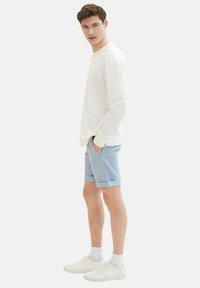 Camisa blanca de manga larga ligera, pantalones cortos azul claro y zapatillas blancas con superficie texturizada. Ajuste casual, con dobladillos enrollados en los pantalones cortos y bolsillos.