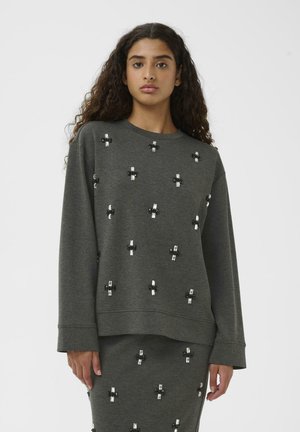 Young woman with curly hair wearing a dark grey long-sleeve top and matching skirt decorated with small black and white cross patterns.