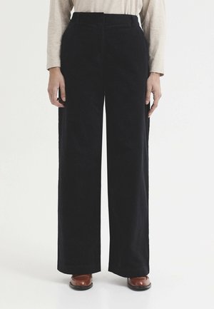 Persona que lleva pantalones negros de tiro alto y pierna ancha, camiseta beige de manga larga y zapatos de cuero marrón, de pie recto frente a un fondo blanco.