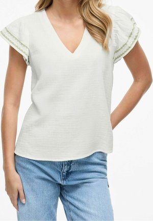 Kvinde iført hvid V-hals bluse med korte flæseærmer med grøn kant, kombineret med lyseblå denimjeans.