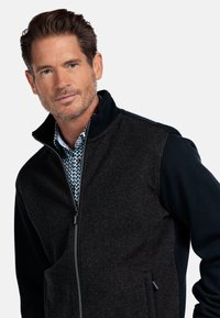 Donker zip-up jack met gestructureerde bovenstof, gladde mouwen en een hoge kraag. Gecombineerd met een bedrukte shirt eronder.