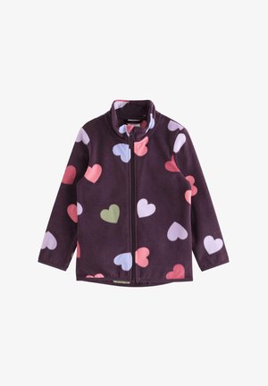 Ung pige iført en lilla hue, lilla jakke med hjerte-mønster, lyserød t-shirt, løstsiddende mørke jeans og pink sneakers, stående med armene langs siden.