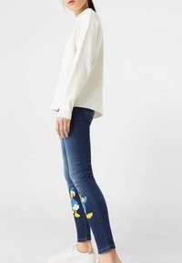 White long-sleeve top, blue skinny jeans with embroidered Donald Duck, and white sneakers. Clean design and casual fit.