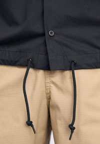 Svart skjorta med knappstängning och dragsko i nederkant, tillsammans med strukturerade beige byxor med ett subtilt rutmönster.