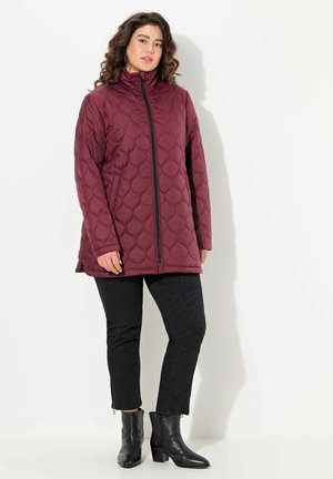 Burgundy quilted long jacket with high collar, front zipper, and side pockets, paired with black patterned pants and black heeled boots.