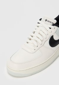 Bijeli tenisice s crnim Nike logom, Gore-Tex oznakom, bijelim vezicama i teksturiranim potplatom na jednostavnoj svijetloj pozadini.