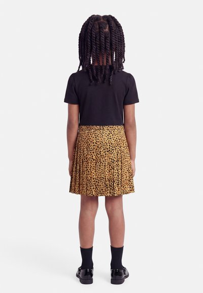 Enfant aux cheveux tressés, vue de dos, portant un haut noir à manches courtes, une jupe plissée à motif léopard, des chaussettes noires et des chaussures noires.
