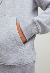 Lys grå sweatshirt med kengurulomme, ribbede mansjetter og et blødt, struktureret stof. Detaljeret syning fremhæver designet.