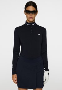 Langärmliges, schwarzes Polohemd mit hohem Kragen und dreiknöpfiger Knopfleiste, kombiniert mit einem dunklen Rock. Accessoiert mit Sonnenbrille und einem weißen Golfhandschuh.