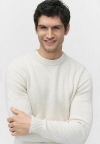 Vit, stickad tröja med ribbad krage och ärmslut. Modellen poserar med armarna i kors och visar en neutral min mot en ljus bakgrund.