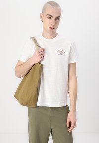 Hvit t-skjorte med korte ermer og teksturert stoff, brun grafikk på brystet, sammen med en beige kanvas totebag. Olivengrønne bukser fullfører antrekket.