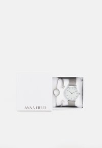 Silver watch with a white face, mesh band, accompanied by a silver bracelet with circular charm and small diamond-like accents.