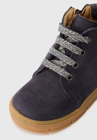 Chaussure en daim bleu foncé avec des lacets texturés gris et blancs et une semelle en caoutchouc beige, présentée sur un fond blanc.