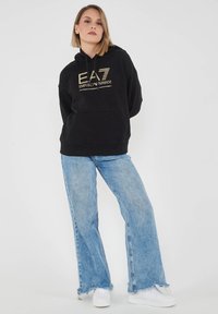 Felpa nera con cappuccio e dettagli del logo in oro, tasca frontale e polsini a coste. Abbinata a jeans a palazzo azzurri chiari e sneaker bianche.
