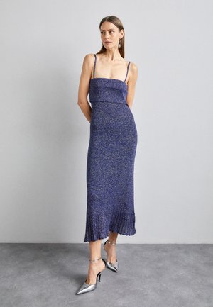 Rochie midi tricotată, de culoare purpurie, cu bretele subțiri, bust ajustat și tiv evazat. Asortată cu pantofi cu toc înalt, argintii, cu vârf ascuțit.