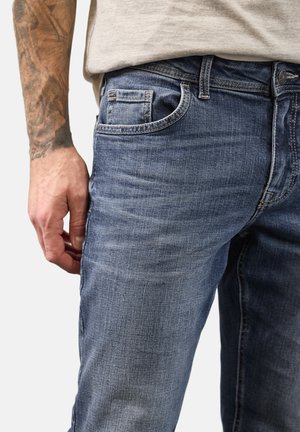 Detailní záběr na tetovanou paži a ruku vedle modrých džínů s viditelným prošíváním a předním kapsou, nošené s světle šedým zastrčeným tričkem.