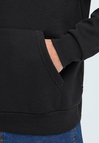 Svart hettegenser med lomme foran, med ribbestrikket mansjetter og hem. Glatt tekstur, brukt sammen med dongri-jeans.