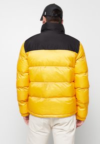Gelber Pufferjacke mit einem schwarzen oberen Abschnitt, ausgestattet mit einem hohen Kragen und einem gesteppten Design. Glatte Textur mit sichtbaren Nähten und Polsterung.