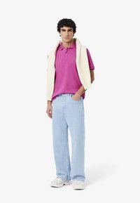 Rosa polo shirt med två knappar, ljusblå baggy jeans och vita sneakers. En krämfärgad stickad tröja är draperad över axlarna.