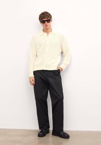 Kardigan w kolorze off-white, o fakturowanym splocie, z kołnierzykiem, z zapięciem na guziki, zestawiony z luźnymi czarnymi spodniami i czarnymi butami.