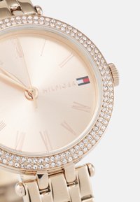 Reloj tono dorado con una correa metálica en rosa dorado, esfera redonda, bisel adornado con cristales y marcas minimalistas en números romanos. Logotipo de "HILFIGER" exhibido.