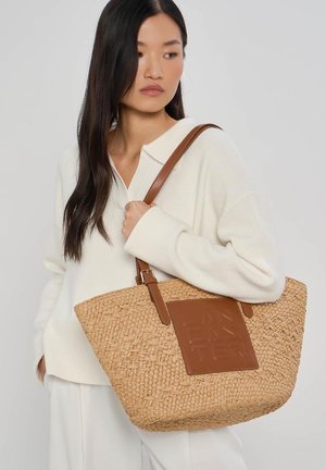 Femme portant un pull blanc côtelé et un pantalon, tenant un grand sac cabas en paille tissée avec des anses en cuir marron et un patch.