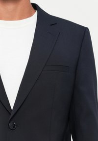Man i svart kavaj över en vit T-shirt med rund hals, som visar övre delen av bröstet och axelområdet.