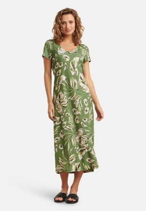 Maxi-Kleid in Grün mit weißen tropischen Blattformen, kurzen Ärmeln, V-Ausschnitt, glattem Stoff und lockerer Passform. Flache schwarze Sandalen.