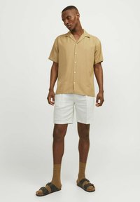 Chemise courte à manches, col, de couleur marron clair avec des boutons blancs, associée à un short blanc. Complétée par des sandales sombres et des chaussettes brunes.