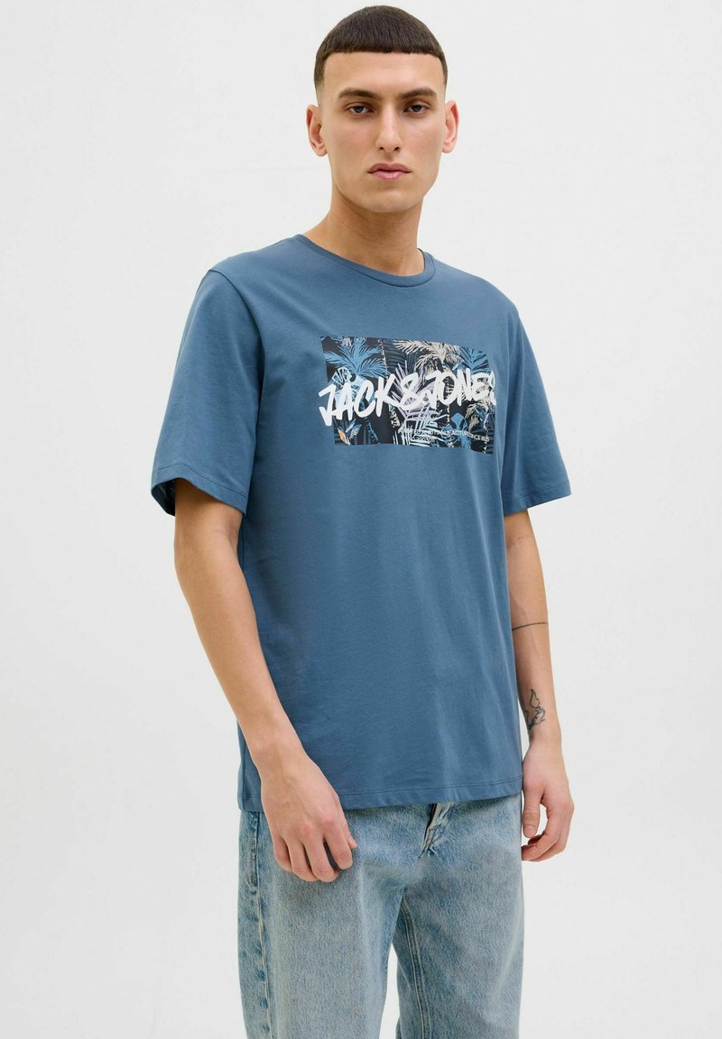 Blaues Baumwoll-T-Shirt mit kurzen Ärmeln, versehen mit einem grafischen Druck von tropischen Blättern und dem Text "JACK & JONES" in Weiß. Minimalistisches Design.