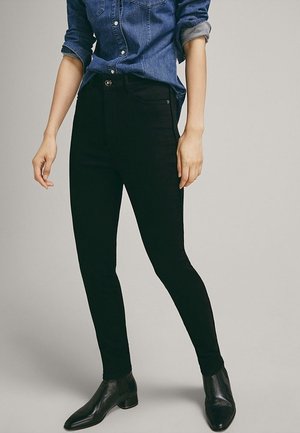 Personne portant un pantalon noir slim, des bottines en cuir noir et une chemise en denim à manches longues retroussées, debout devant un fond uni.