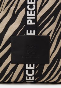 Sac en toile avec un motif de rayures zèbre beige et noir, une étiquette logo en cuir noir et une large bandoulière noire avec des lettres blanches.