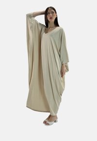 Beige losse kaftan met een V-hals en brede mouwen, gemaakt van zachte stof. Het model draagt witte open-teenhakken.