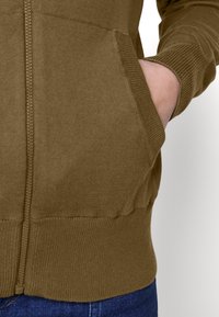 Oliven grøn zip-up hættetrøje med ribbede manchetter og talje. Har sidelommer. Materialet virker blødt og tekstureret.