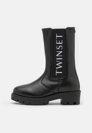 Bota negra de cuero hasta la mitad de la pantorrilla con suela y tacón gruesos, panel lateral elástico que muestra el texto vertical "TWINSET" en blanco.