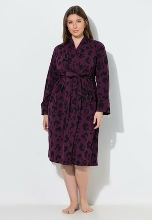 Kvinde står barfodet iført en knælængde lilla kimono med sorte leopardpletter og et bundet bælte mod en ensartet lys baggrund.
