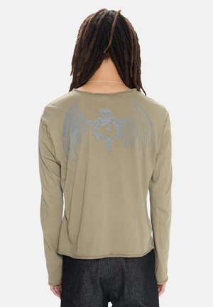 Personne aux longues dreadlocks portant un t-shirt à manches longues vert olive avec un emblème ailé bleu délavé dans le dos et un pantalon foncé.