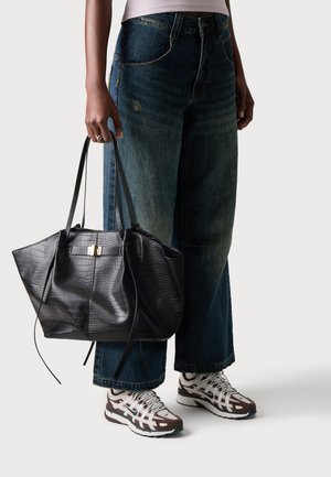 Personne portant un jean large foncé et des baskets blanc-marron tenant un grand sac à main en cuir noir texturé par ses anses.