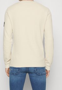 Ljust beige långärmad skjorta med en texturerad tyg, rund halsringning och en svart accent på vänster ärm. Kombinerad med blå jeans.